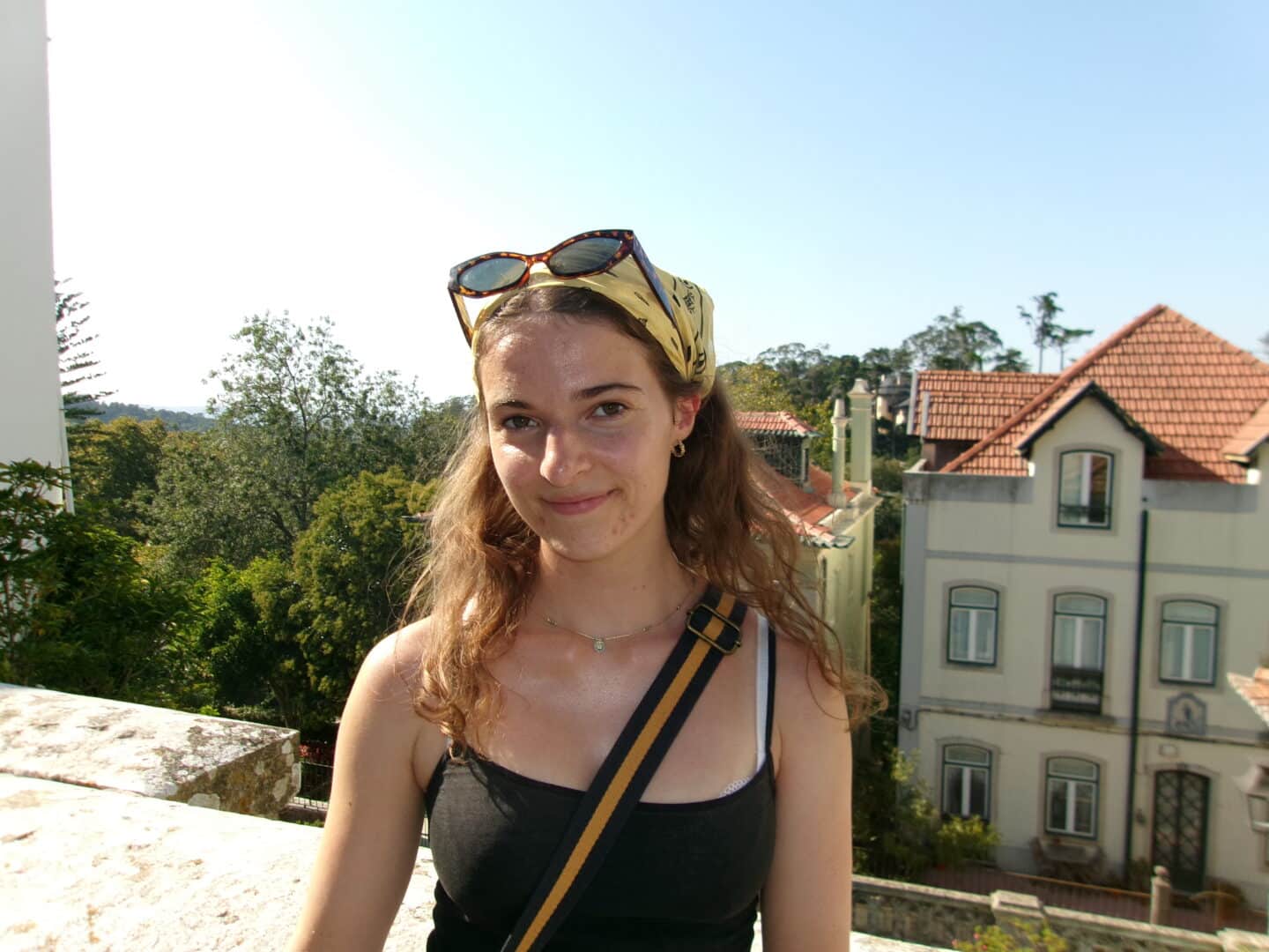 A young person standing outside in a summery scene with large, old buildings and trees in the background.