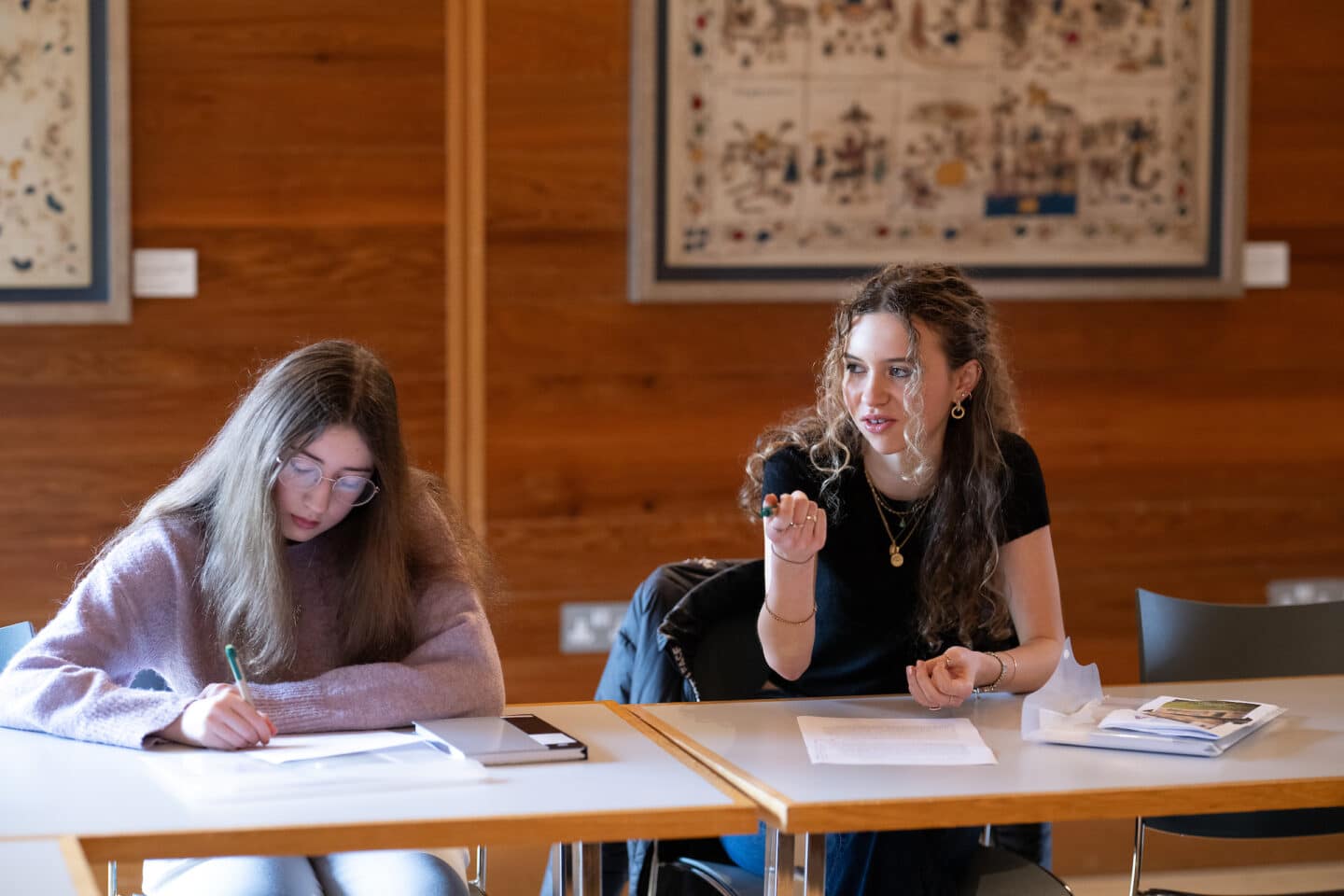 Two students are sitting at a desk in class, one is writing and one is talking