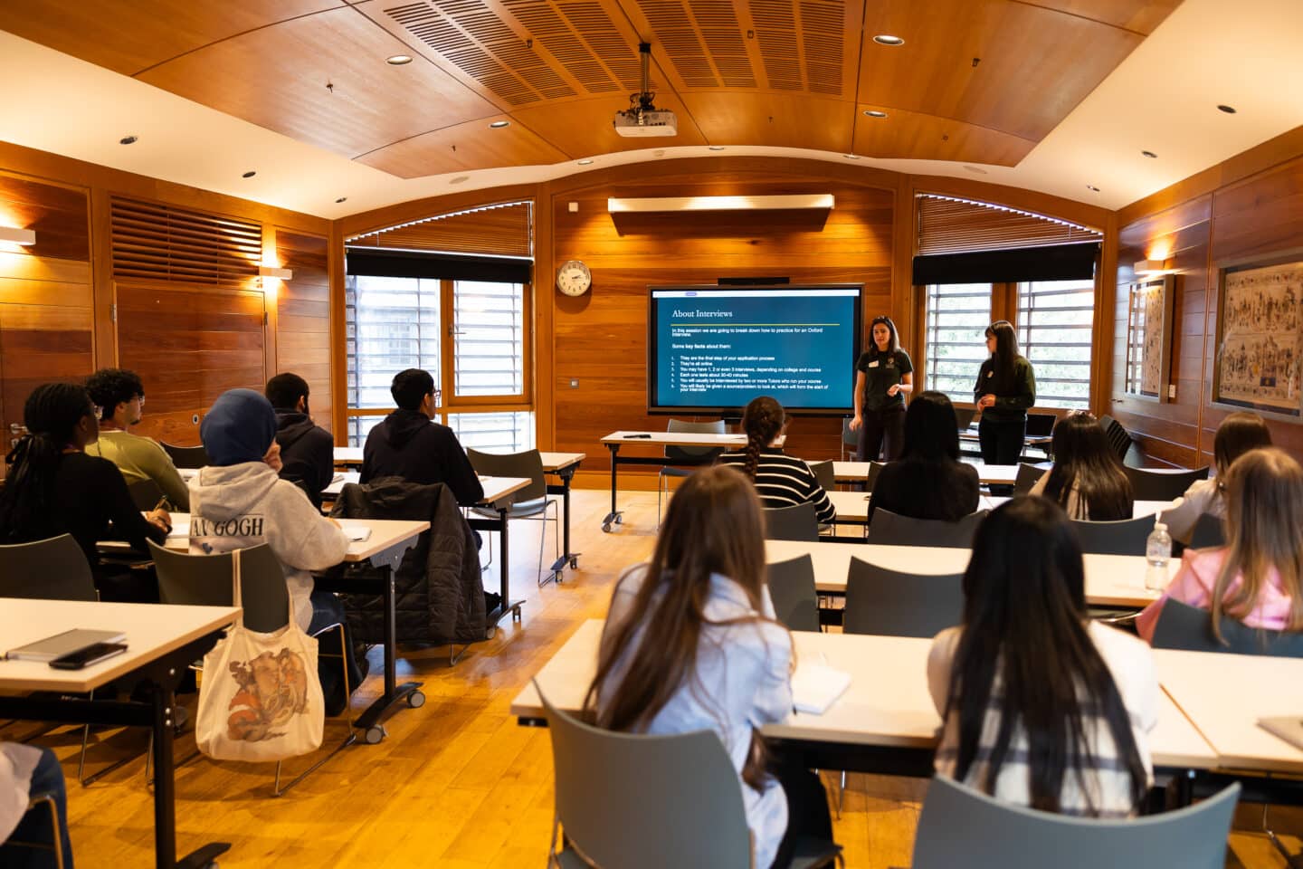 a group of students listening to a presentation from two young adults in a large wooden classroom.