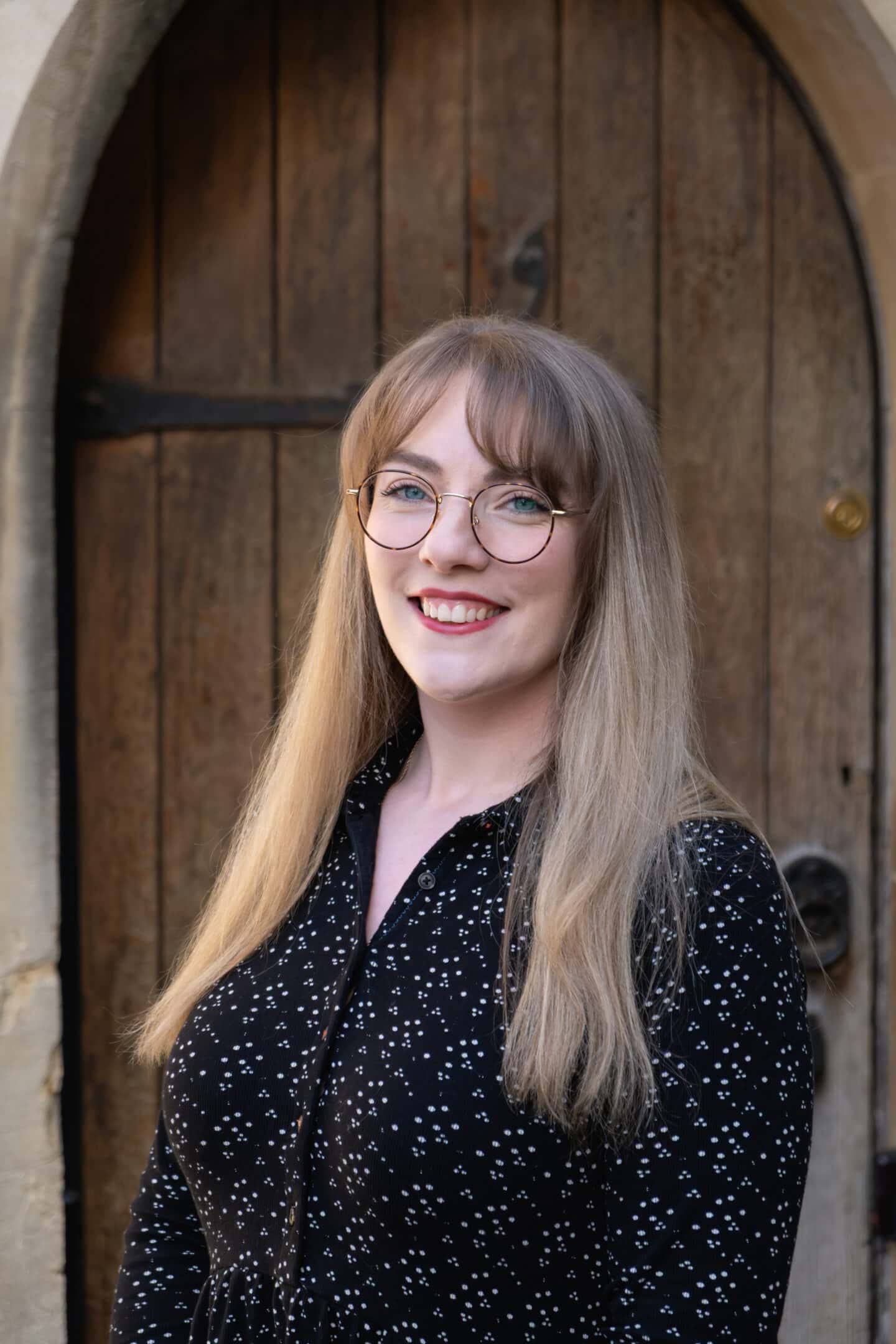 A woman with glasses standing outside a building with an old wooden door