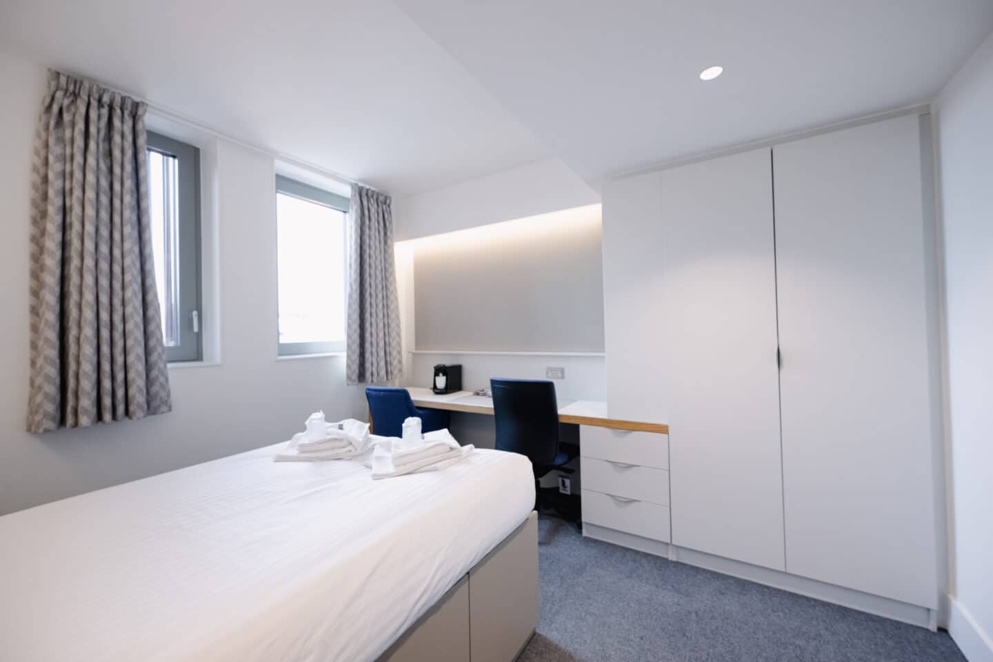 A student bedroom with a desk, wardrobe, bed with underbed storage and windows