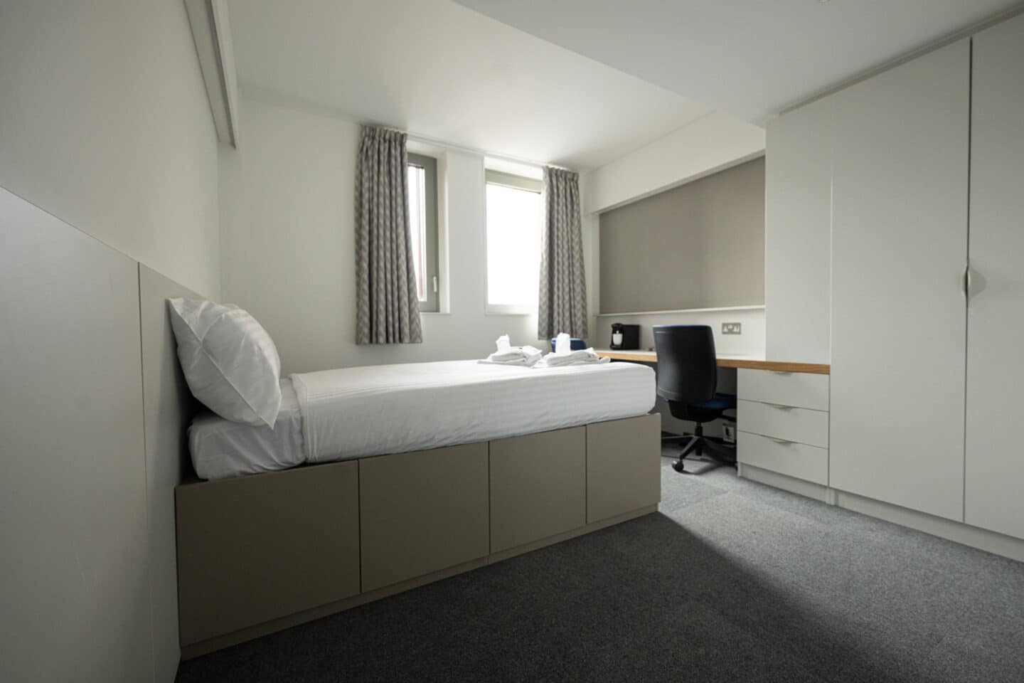A student bedroom with a desk, wardrobe, bed with underbed storage and windows