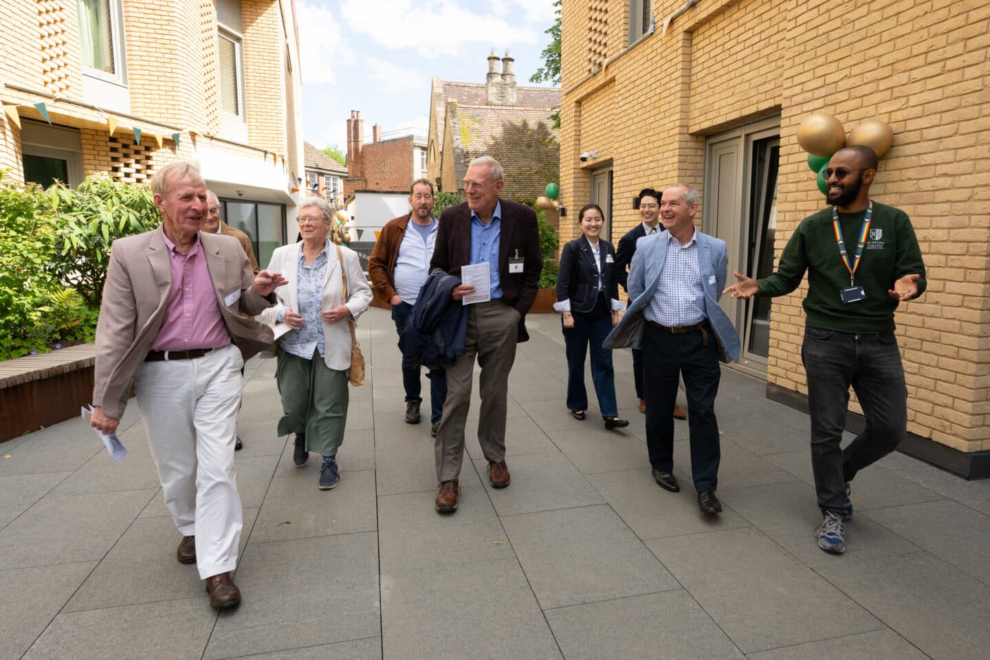 Eight people walking with a tour guide between two light-coloured brick buildings decorated with bunting and balloons