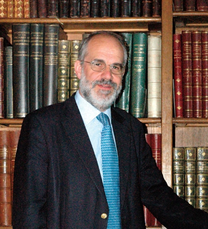 George Brooke wearing a suit and tie and standing in front of a bookshelf with old leather-bound books