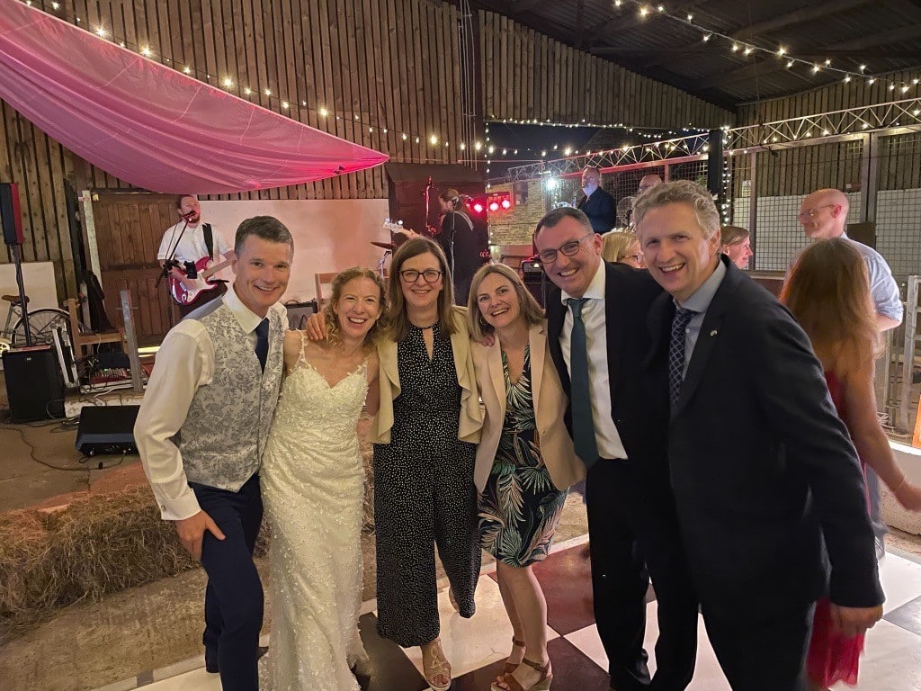 six adults, three women and three men, at a wedding. One couple is the bride and groom.
