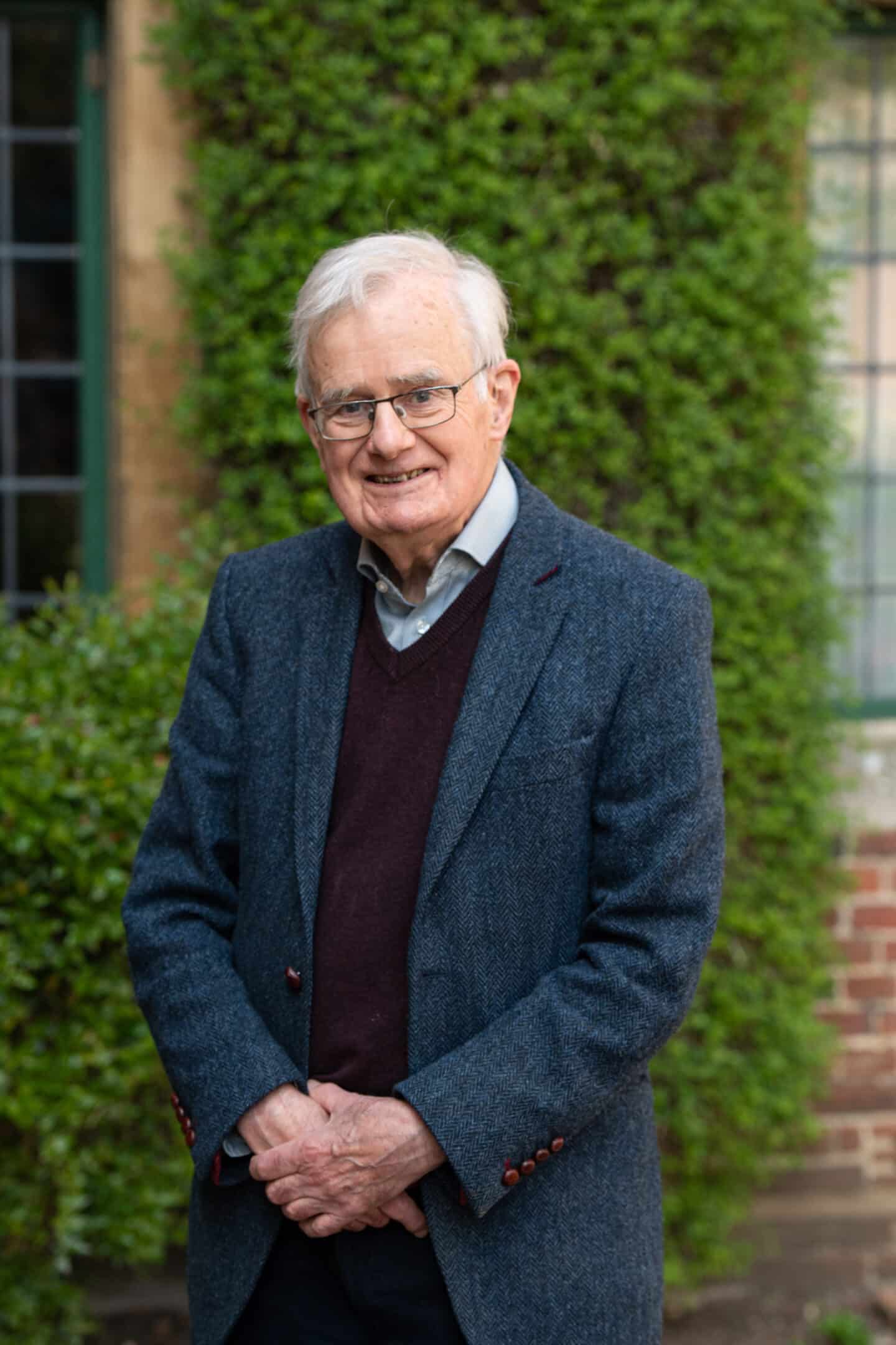 A headshot of a man wearing a tweed coat, sweater, and glasses