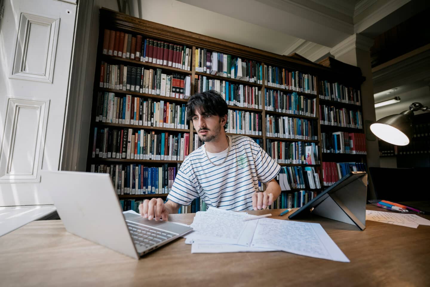 A student wearing a striped T-shirt is sitting at a desk in the library, in front of shelves full of books. They are looking at something on a silver laptop, and there are a pile of papers filled with handwritten work in front of them.
