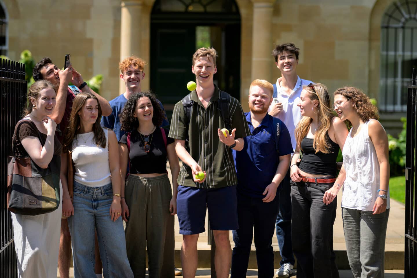 group of students standing outside on a sunny day watching one student juggle three tennis balls