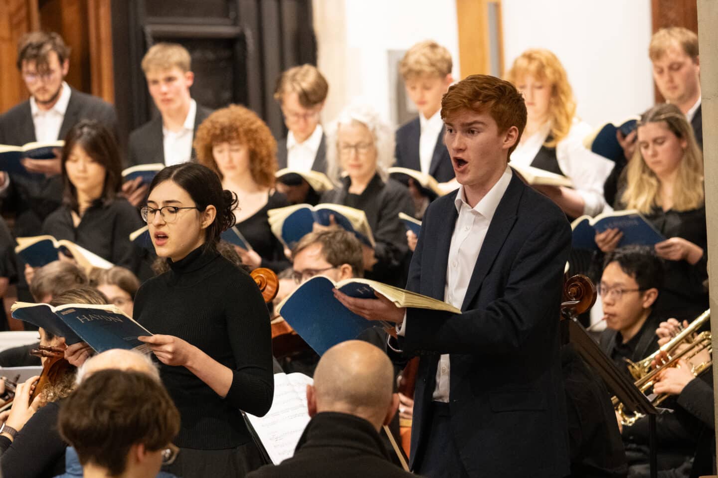 Two student soloists holding sheet music and singing, with a choir and orchestra in the background