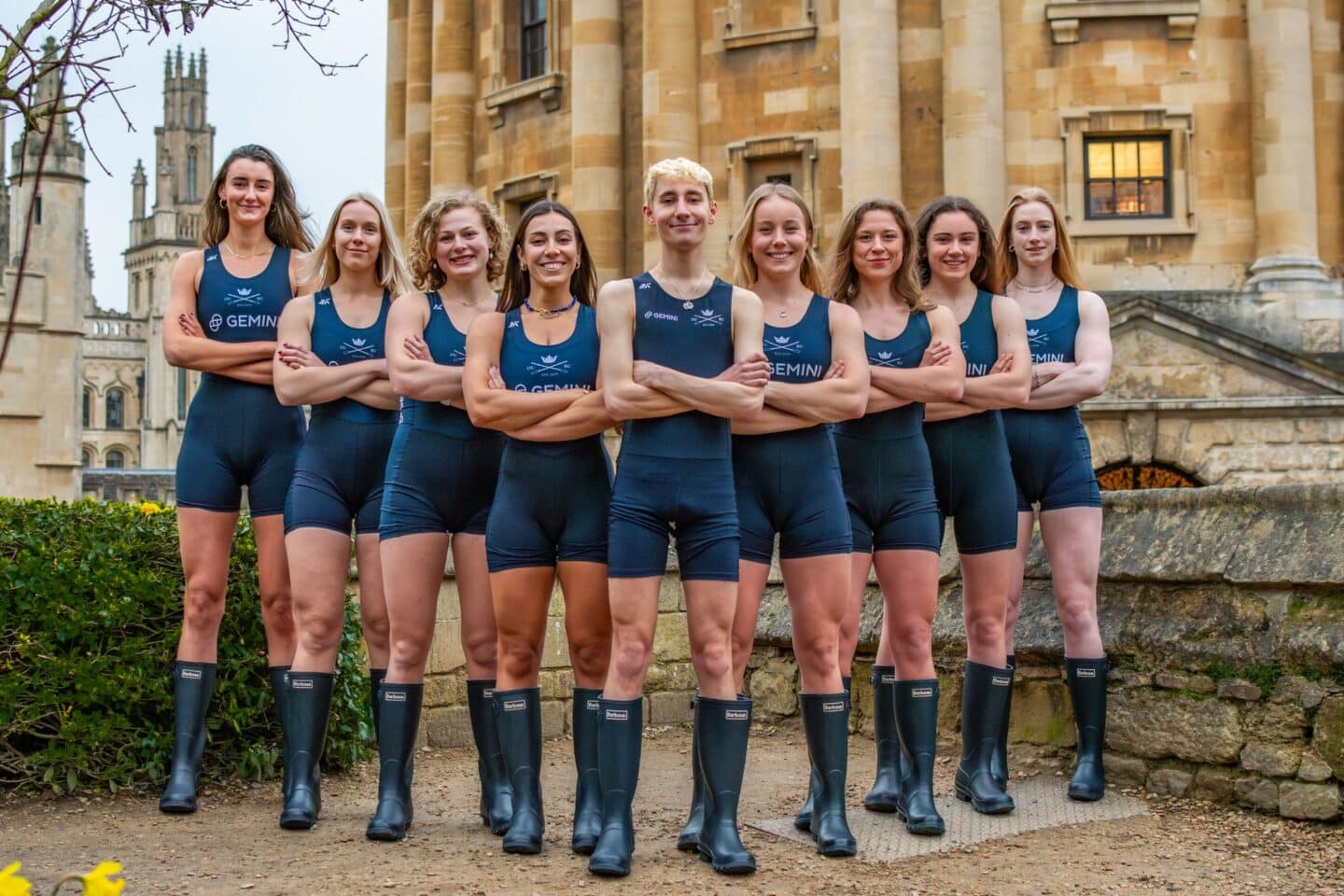 Nine people wearing rowing gear, standing with arms crossed with an old stone building in the background