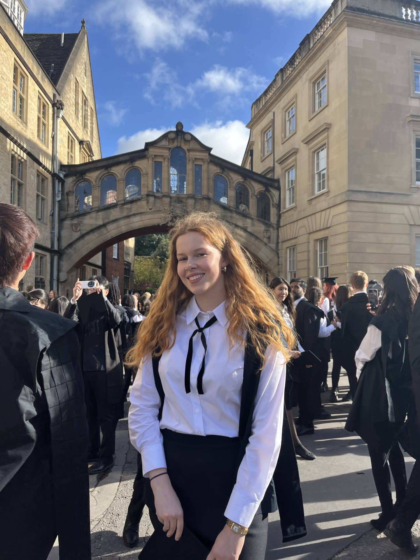 A smiling student in sub fusc in front of a busy street in Oxford under the Hertford bridge
