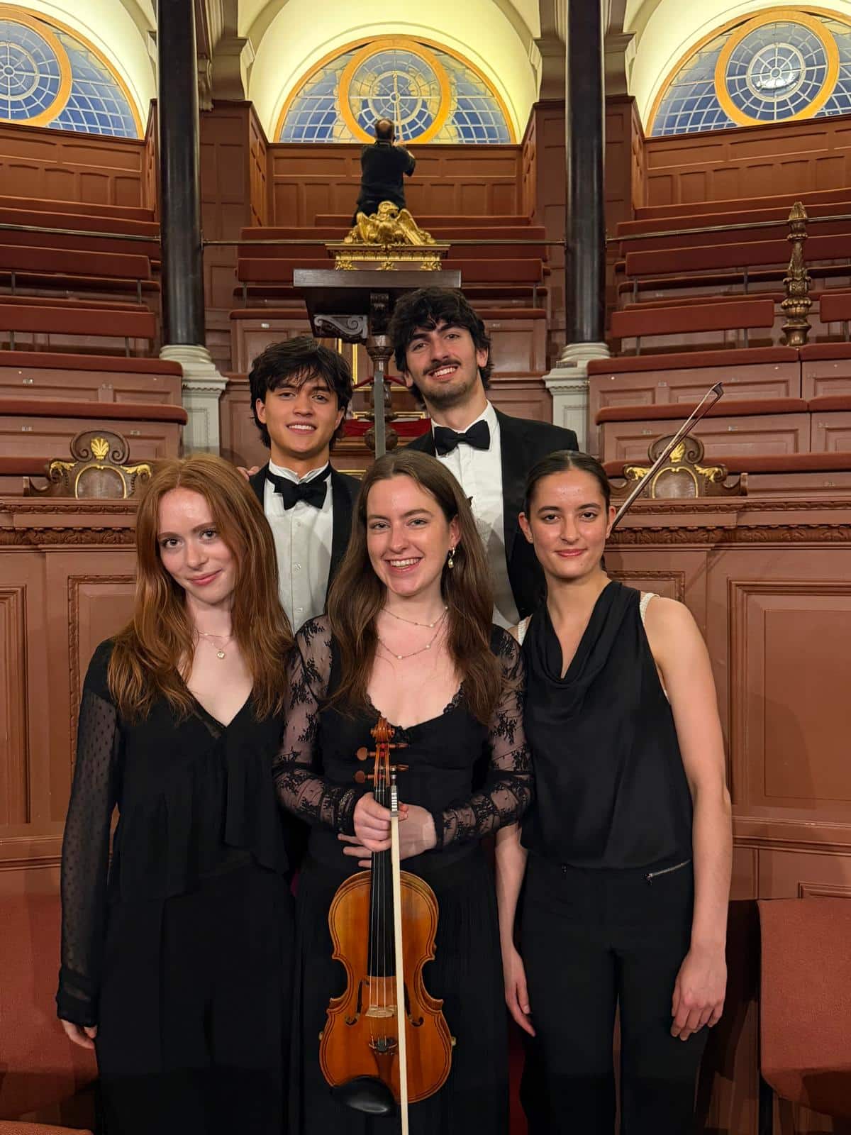 Five students in formal attire in an old building with painted wooden panelling.