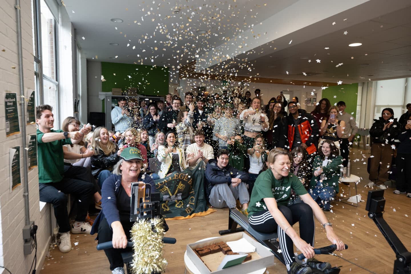 A large group of people throwing confetti as two people on erg machines row