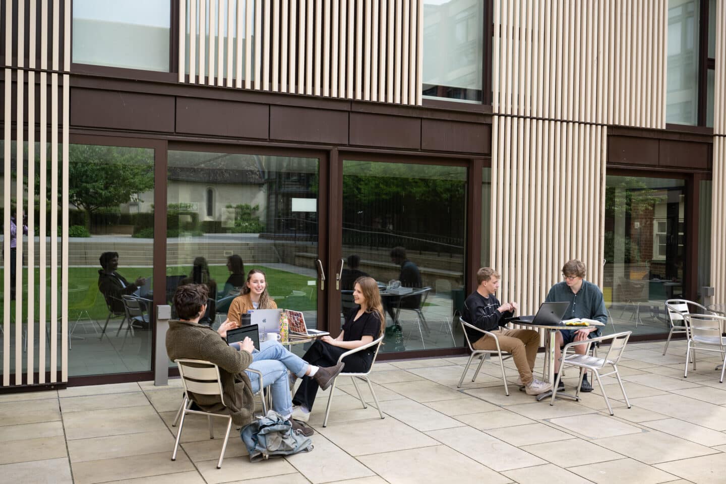 two groups of young people sat at two tables outside a modern building with large windows.