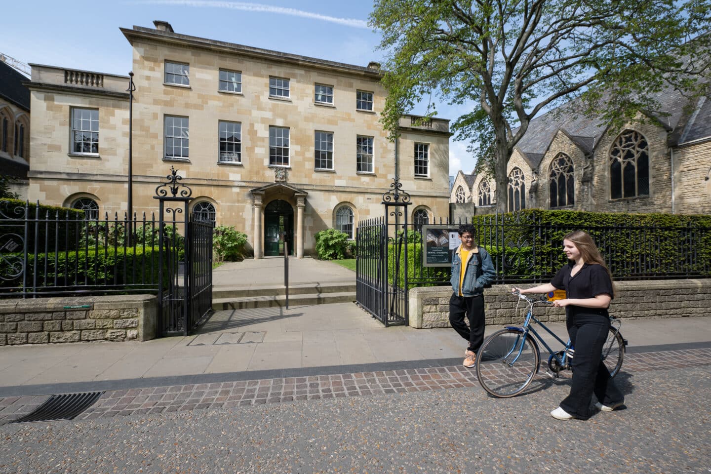 An outdoor scene with large and old limestone buildings. Two students are walking together, and one of the students is walking their bicycle.