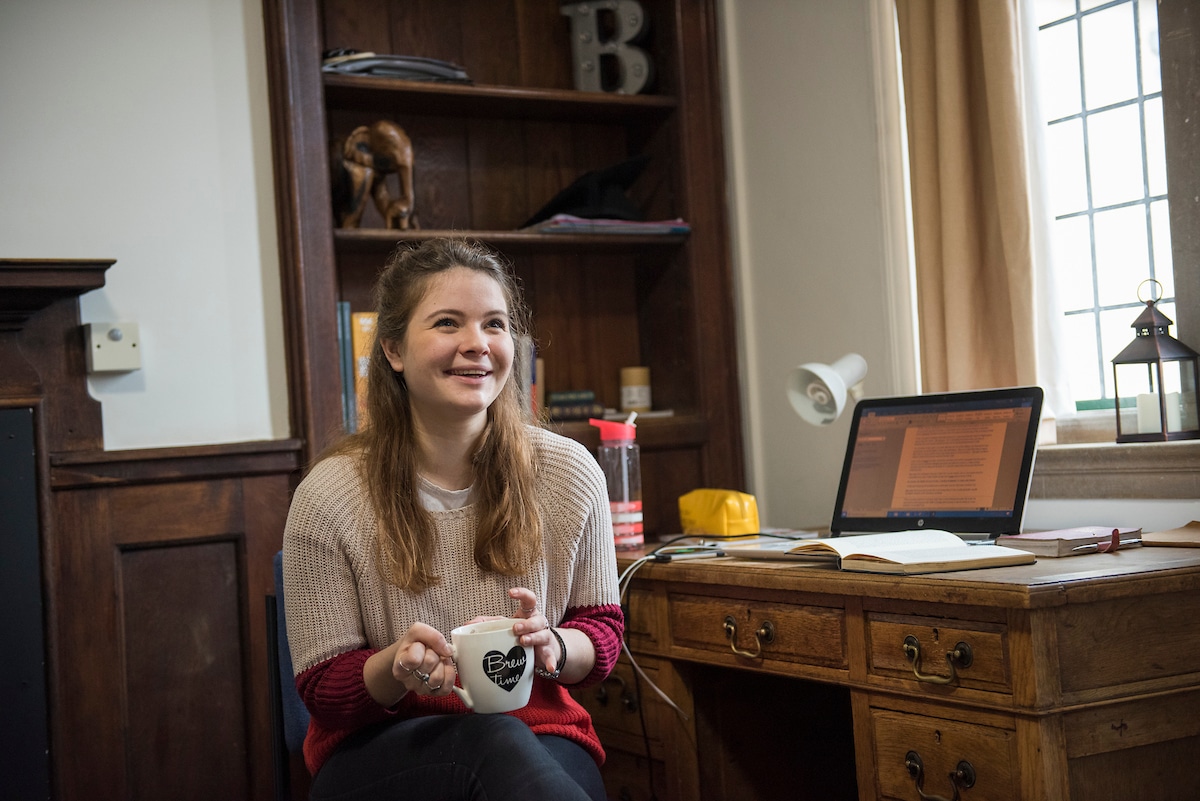 student holding a cup of tea by a desk with a computer and notebook.