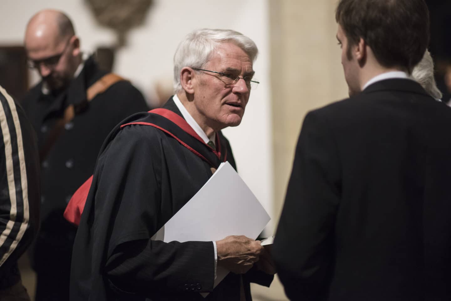 A man in academic regalia wearing glasses and holding a booklet