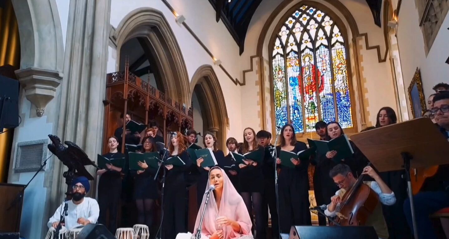 A singer sat on a stage in a chapel, surrounded by musicians and a choir.