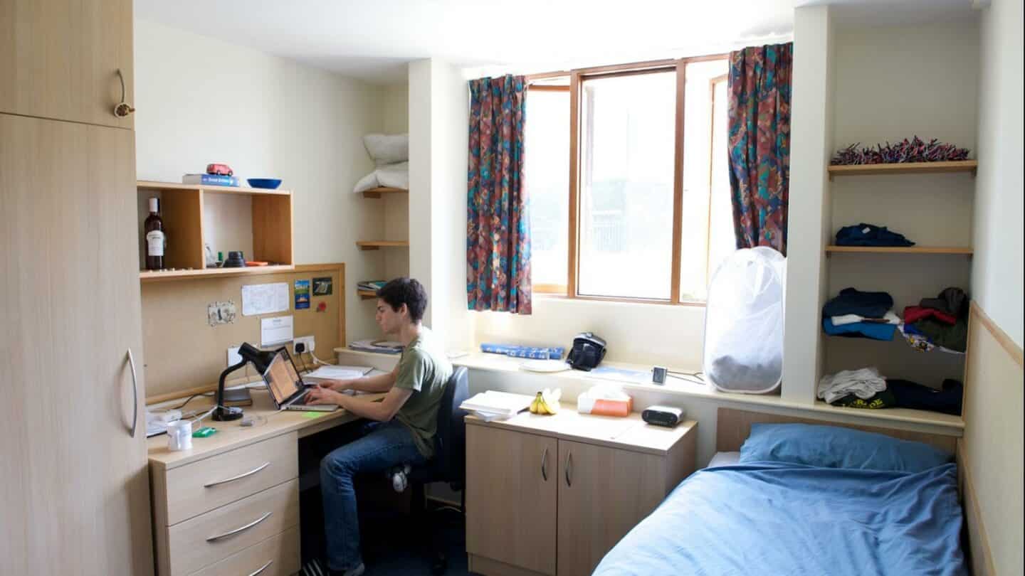 a student sitting in a room with a desk, bed and cupboards