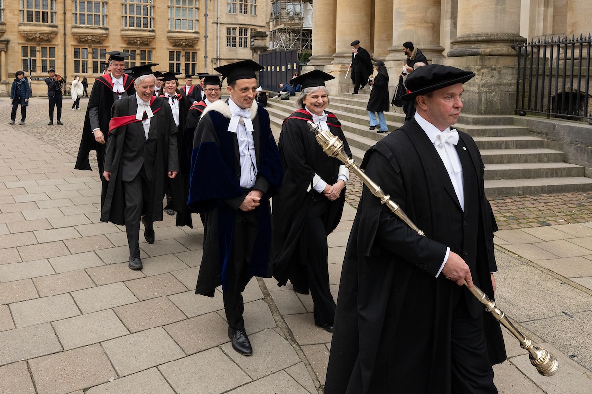 An academic procession taking place on a stone-paved streed, with the group wearing academic gowns. Onlookers are taking photos.