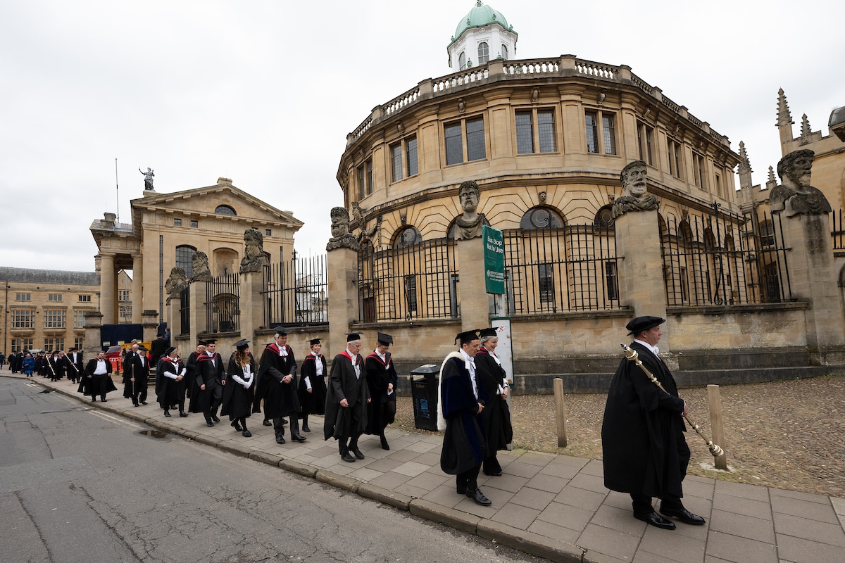 An academic procession taking place on a stone-paved streed, with the group wearing academic gowns. Onlookers are taking photos.