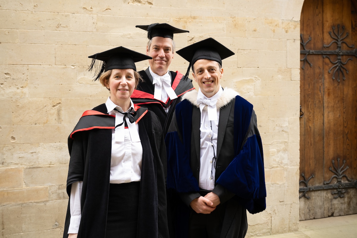 Three people in formal academic dress standing in front of an old limestone building.