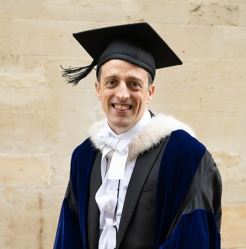 A man wearing an academic gown and cap and smiling