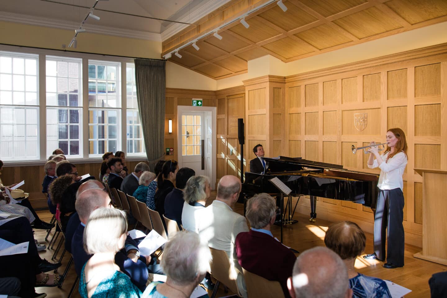 A man playing a small grand piano and a woman playing the trumpet in a wood-paneled room filled.