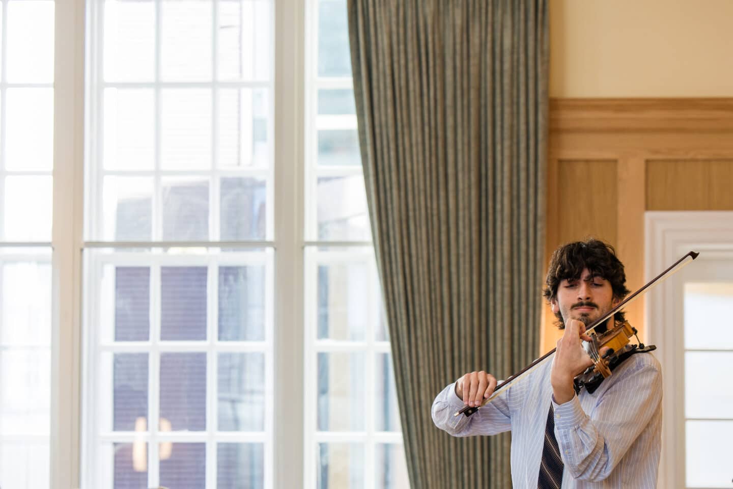 A young man plays the violin in a wood-panelled room with large windows and curtains.