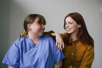 Two women, one in causal wear and one in nurse scrubs, sitting and smiling at one another in a posed photo.